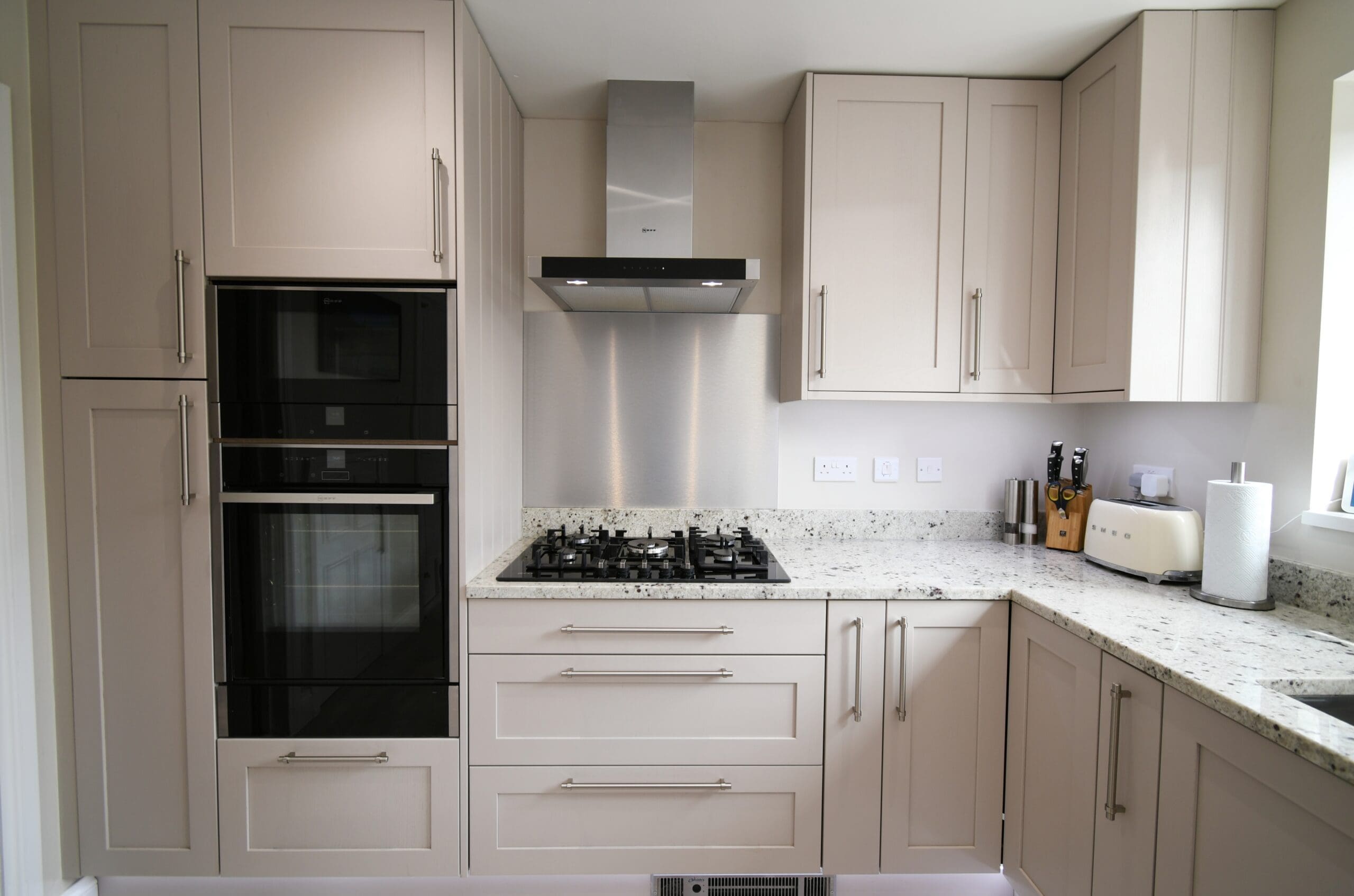 Image of a modern Cashmere Skinny Shaker kitchen featuring sleek, minimalist design. The kitchen includes Neff Slide & Hide ovens seamlessly integrated into the cabinetry, a gas hob with a wall-mounted extractor fan above, and units that extend all the way to the ceiling, providing a clean and sophisticated look